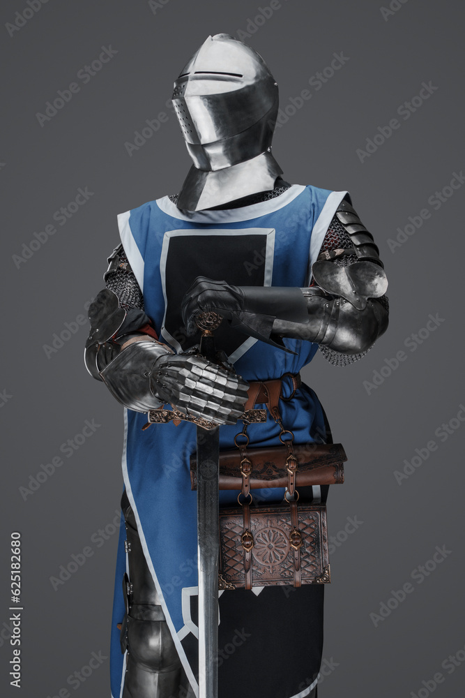 Fototapeta premium Medieval guard dressed in armor and blue surcoat holding a sword, standing in statue pose, against a gray background