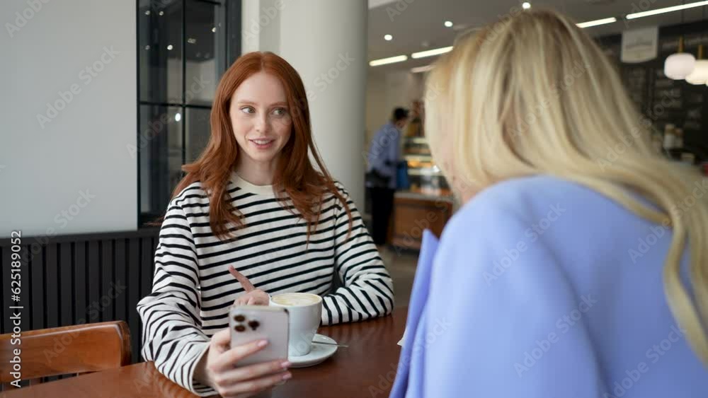 Back view of two young beautiful women friends sitting in cafe indoors, looking at phone and talking, laughing. Two pretty females girlfriends using social media on smartphone in coffee shop. Back view of two young beautiful women friends sitting in cafe indoors, looking at phone and talking, laughing. Two pretty females girlfriends using social media on smartphone in coffee shop.