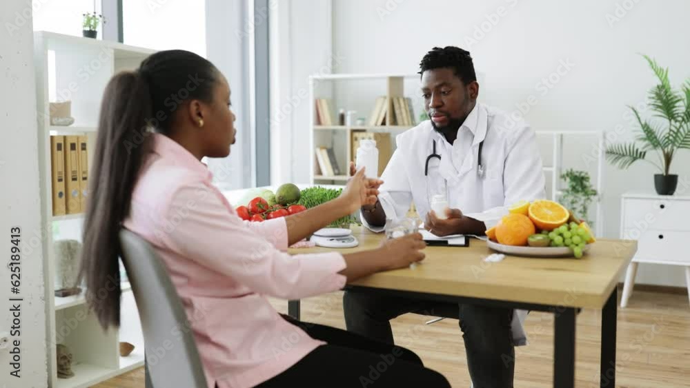 Friendly multicultural man in lab coat holding medicine containers in hands while sitting at writing desk during consultation. Confident expert in nutrition advising on food supplements in meal plan. Friendly multicultural man in lab coat holding medicine containers in hands while sitting at writing desk during consultation. Confident expert in nutrition advising on food supplements in meal plan.