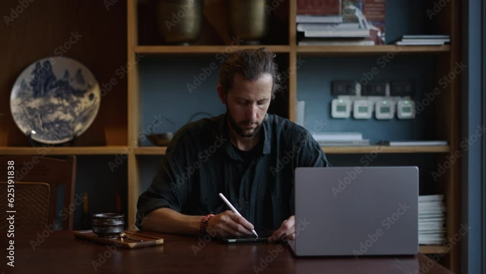 Caucasian designer writes something on his tablet while looking at his laptop Caucasian designer writes something on his tablet while looking at his laptop