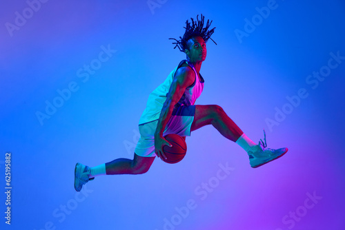 Foto Slam dunk Ambitious young man, basketball player in minions, playing basketball against gradient blue background in neon lights