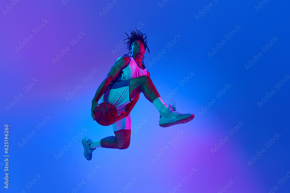Slam dunk Ambitious young man, basketball player in minions, playing basketball against gradient blue background in neon lights. Concept of professional sport, competition, hobby, competition, ad Slam dunk Ambitious young man, basketball player in minions, playing basketball against gradient blue background in neon lights. Concept of professional sport, competition, hobby, competition, ad