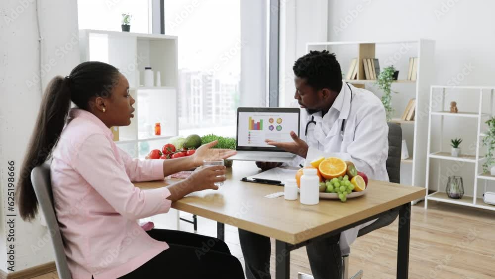 African american doctor and patient having appointment in hospital cabinet and discussing woman's treatment. Long haired lady holding glass of water while male specialist pointing at graphs on laptop. African american doctor and patient having appointment in hospital cabinet and discussing woman's treatment. Long haired lady holding glass of water while male specialist pointing at graphs on laptop.