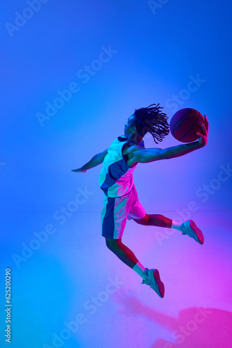 Full-length image of young professional basketball player in motion, training, playing over gradient blue background in neon lights. Concept of professional sport, competition, game, competition, ad