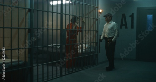 Canvas-taulu Jailer with police baton stands near prison cell, talks to female prisoner in orange uniform through metal bars