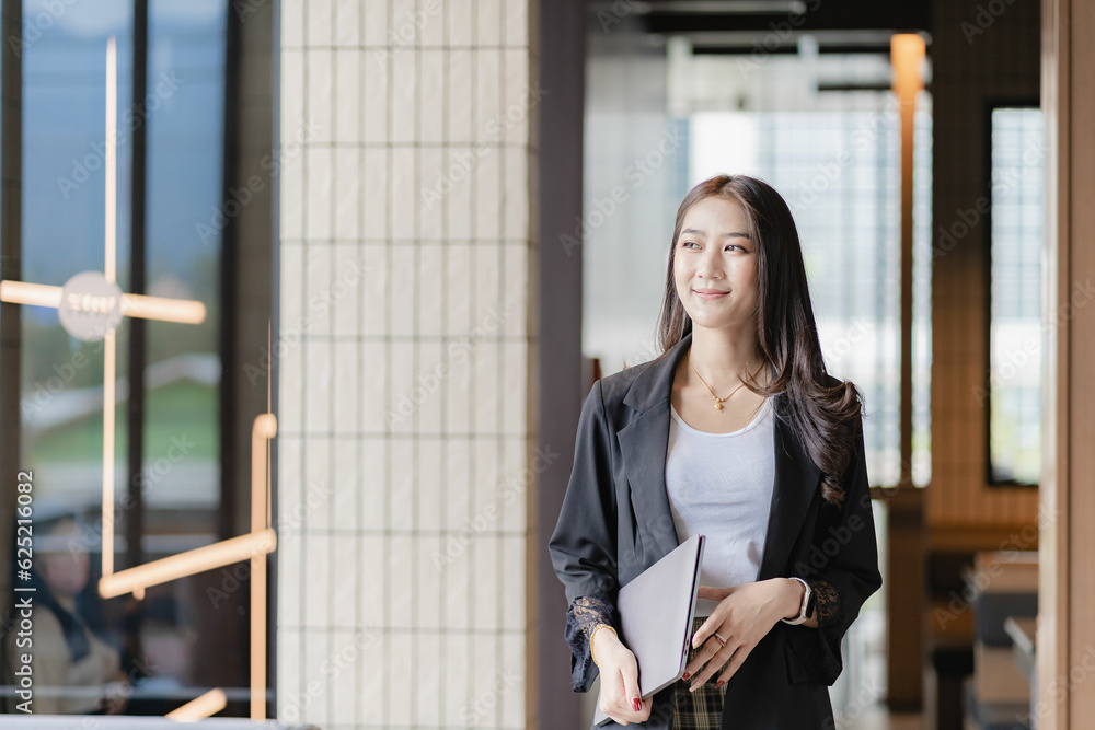 Fototapeta premium Beautiful Asian businesswoman holding a laptop computer Walking smiling while walking into the office