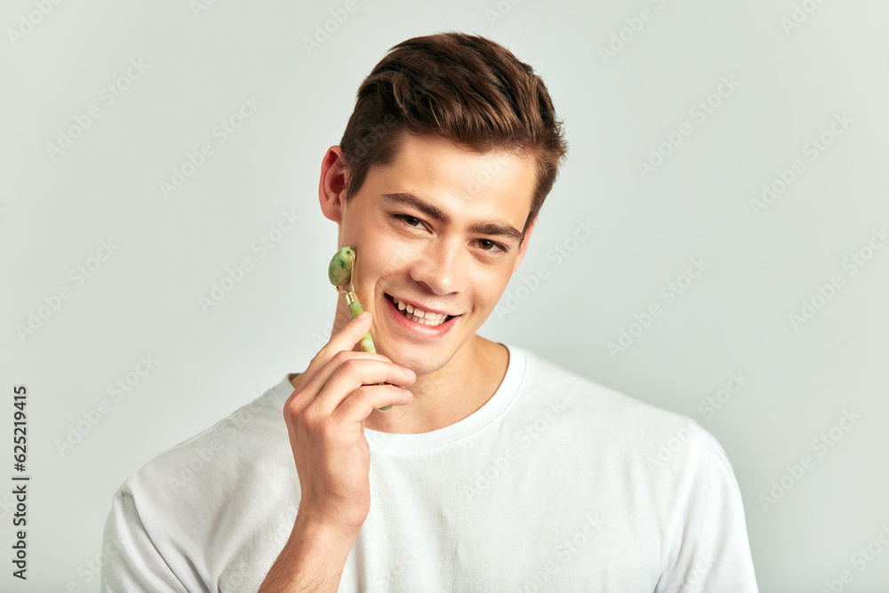 Young handsome smiling brown haired guy using jade de-puffing face roller in studio to keep his skin soft and smooth. Caucasian millennial man maintains his natural beauty with a skincare routine. Young handsome smiling brown haired guy using jade de-puffing face roller in studio to keep his skin soft and smooth. Caucasian millennial man maintains his natural beauty with a skincare routine.