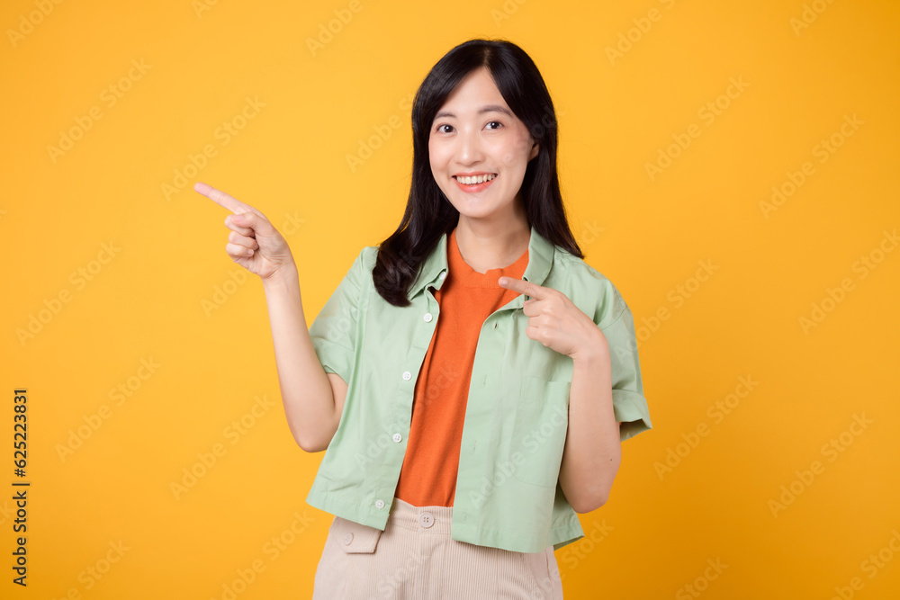 young Asian woman in her 30s wearing a green shirt on an orange shirt, pointing fingers to free copy space. Explore the concept of discount shopping promotion with eye-catching image. young Asian woman in her 30s wearing a green shirt on an orange shirt, pointing fingers to free copy space. Explore the concept of discount shopping promotion with eye-catching image.