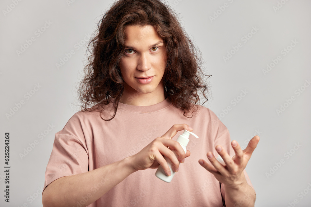 Young handsome long haired guy putting facial wash on his fingers, preparing to apply beauty product on his face. Caucasian man with brown hair uses hydrating skin cleanser in his skincare routine. Young handsome long haired guy putting facial wash on his fingers, preparing to apply beauty product on his face. Caucasian man with brown hair uses hydrating skin cleanser in his skincare routine.