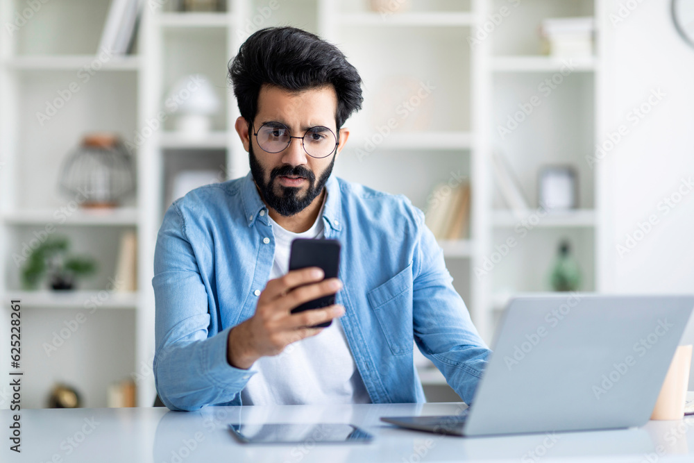 Stressed indian male freelancer looking at smartphone screen in home office Stressed indian male freelancer looking at smartphone screen in home office