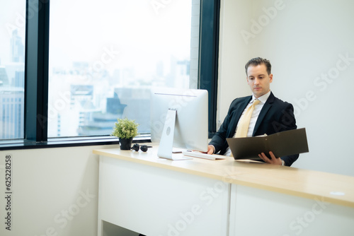 Business man in formal dressed looking at hard copy file seriously while using computer at desk in highg building office  beside glass window with building view. photo