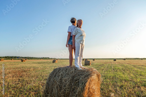 Smiling elderly and young women stand on a roll of hay in a field at sunset. Harvesting, labor and freedom.