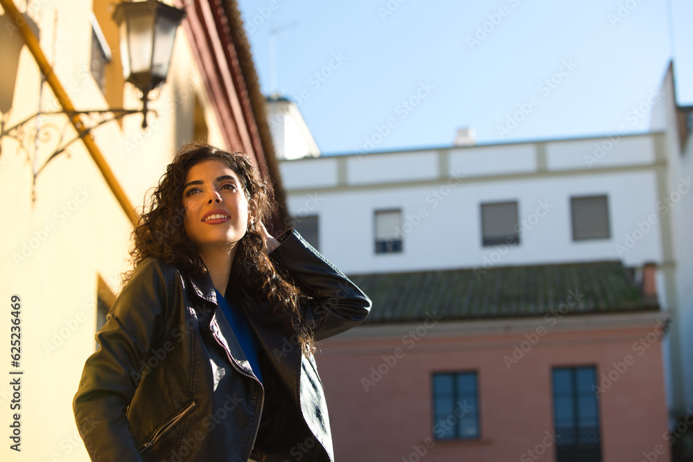 Fototapeta premium Young and beautiful Hispanic brunette woman with curly hair and black leather clothes is sitting on a railing in the street. The woman is happy and smiling.