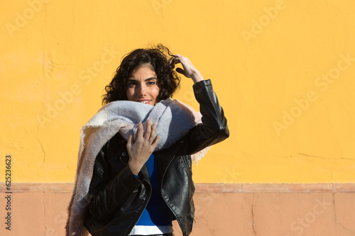 pretty young woman with curly brunette hair against a yellow background is dressed in winter clothes and wearing a scarf to protect herself from the cold. The woman is happy and having fun.