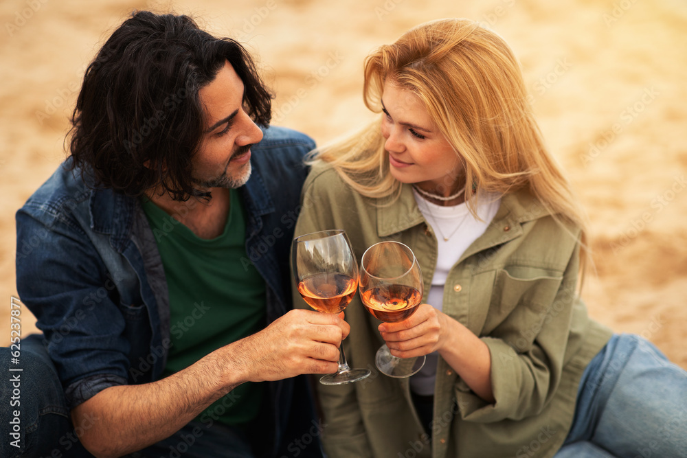 Romantic young couple toasting each other with wine while sitting on beach Romantic young couple toasting each other with wine while sitting on beach