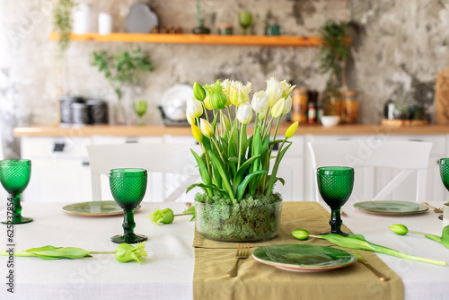 Table festive setting with tulips flowers and stabilization moss in glass vase. Wedding table decoration. Beautiful served table with runner on table, wineglass and plates in living room	 photo