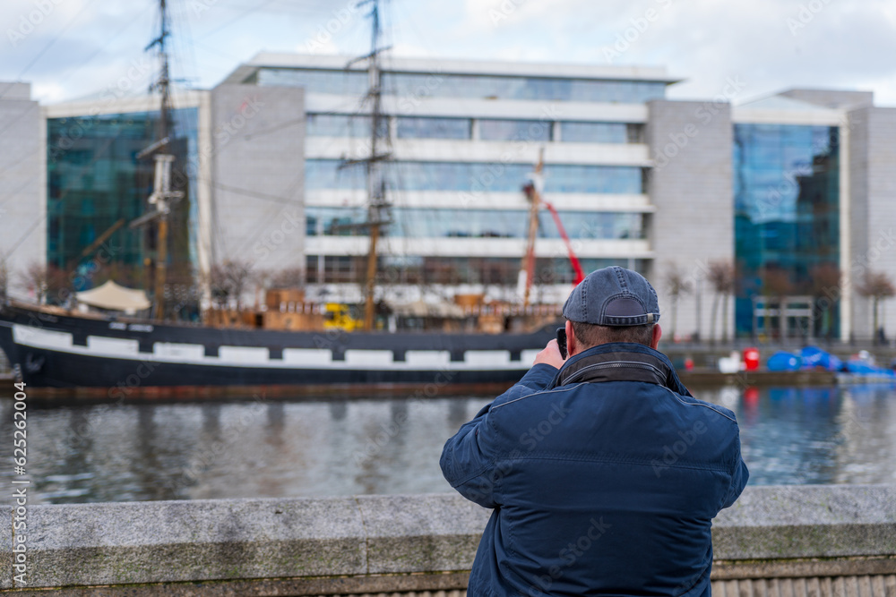 Man wearing a flat cap and blue jacket takes a picture with his smartphone of a sailing ship across the riverbank of the Liffey in Dublin, Ireland Man wearing a flat cap and blue jacket takes a picture with his smartphone of a sailing ship across the riverbank of the Liffey in Dublin, Ireland