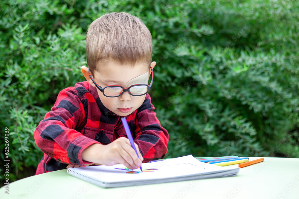 A little boy draws in the garden (outdoors). He wears glasses and an eye patch (occluder) to prevent amblyopia and strabismus (squint, lazy eye). The school students vision diseases problem. A little boy draws in the garden (outdoors). He wears glasses and an eye patch (occluder) to prevent amblyopia and strabismus (squint, lazy eye). The school students vision diseases problem.