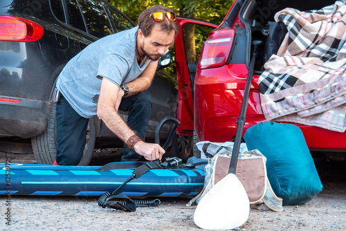 Young bearded guy inflates the sup board with a pump near his car. One shot in series of camping
