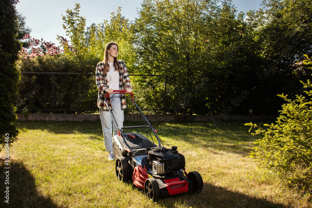 Female gardener working in summer, cutting grass in backyard. Concept of gardening, work, nature. Housework, gardening and country life. Home garden grass cutting woman mowing with lawn mower.