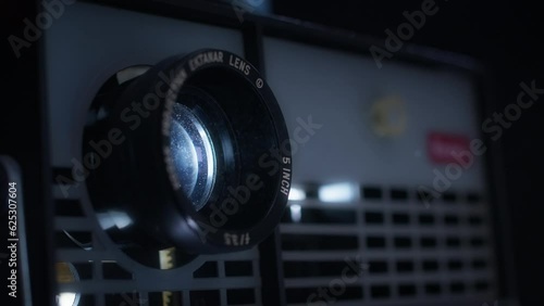 A vintage carousel photographic slide projector advances through transparencies. The projector projects light from its lens, highlighting motes of dust in the air.