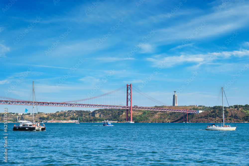 View of 25 de Abril Bridge famous tourist landmark over Tagus river, Christ the King monument and a tourist yacht boat. Lisbon, Portugal