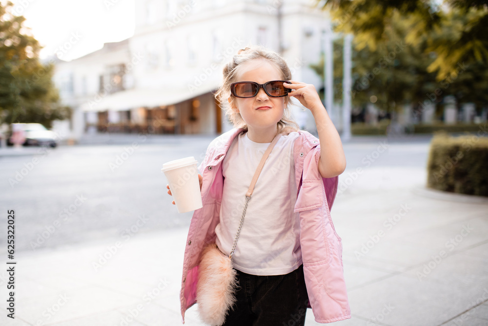 Cute kid girl 4-5 year old wear sun glasses and trendy casual clothes holding coffee cup having fun at city street over sunset outdoor. Portrait of cheerful and happy child. Summer season.