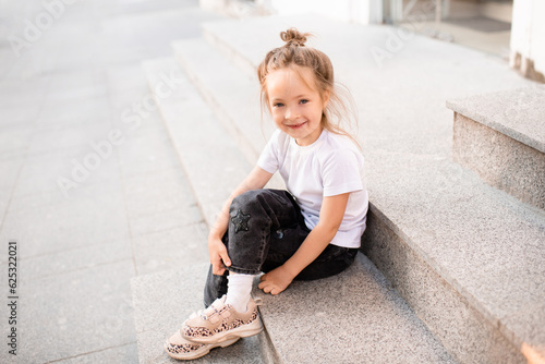 Cute smiling kid girl 4-5 year old wear casual clothes sitting on stairs at city street outdoor. Look at camera. Summer season. Childhood.