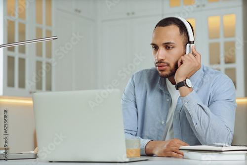 Young man with headphones working on laptop at desk in kitchen. Home office