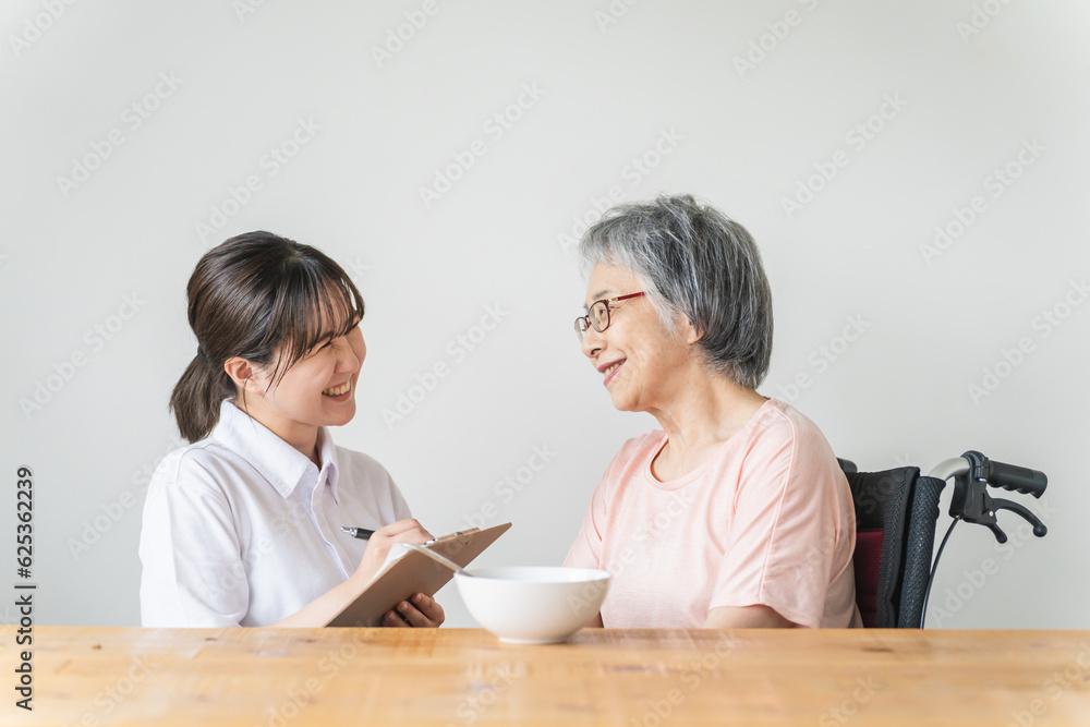 老人ホーム・家で食事する笑顔の高齢者女性と食事介助する管理栄養士・介護士・ヘルパーのアジア人女性
