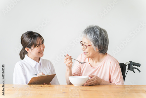 老人ホーム・家で食事する笑顔の高齢者女性と食事介助する管理栄養士・介護士・ヘルパーのアジア人女性
 photo