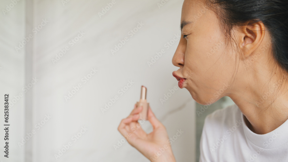 Young Asian happy woman make up looking at her reflection in mirror apply lip balm on her lips in bathroom. Lifestyle and people, Lips Protection, Natural Skin Care Cosmetics, Morning routine concept. Young Asian happy woman make up looking at her reflection in mirror apply lip balm on her lips in bathroom. Lifestyle and people, Lips Protection, Natural Skin Care Cosmetics, Morning routine concept.