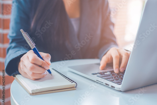 Woman hand is writing on a notepad with a pen and have a laptop computer on the desk in the office.