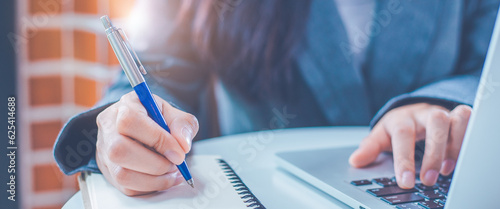Woman hand is writing on a notepad with a pen and have a laptop computer on the desk in the office.
