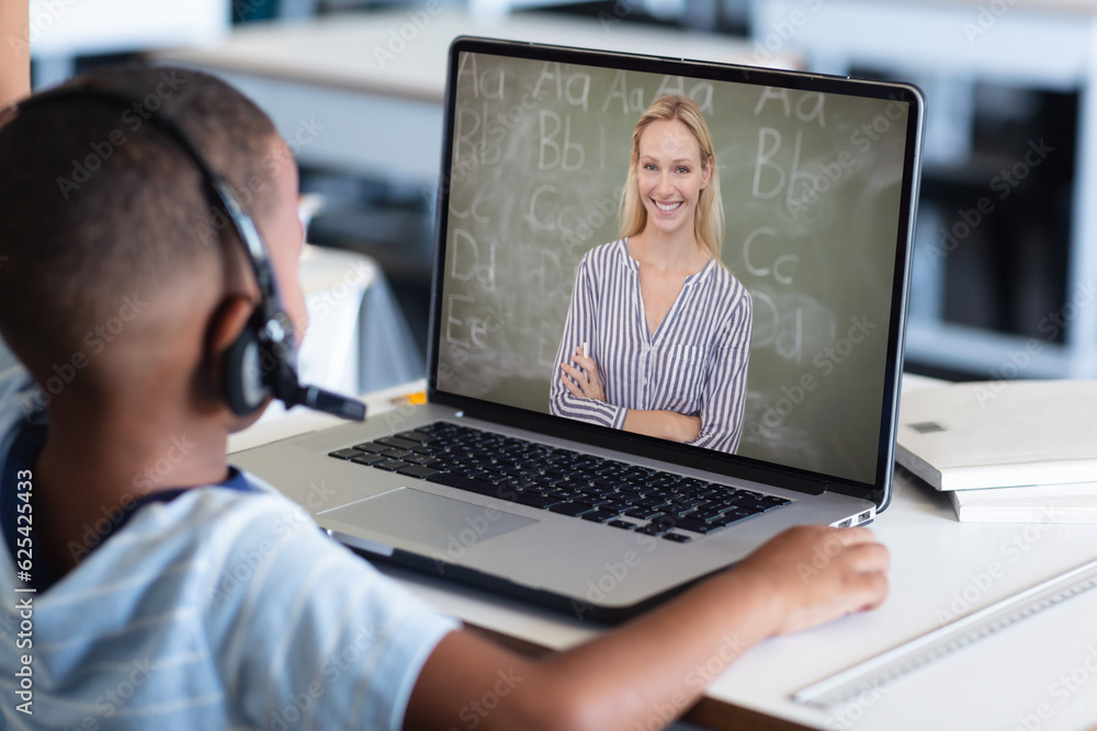 African american boy wearing headphones and listening to female teacher over video call on laptop