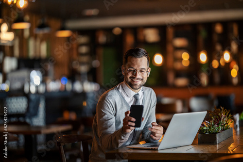 A happy young elegant businessman in smart casual is sitting in a coffee shop with his laptop and smiling at the phone.