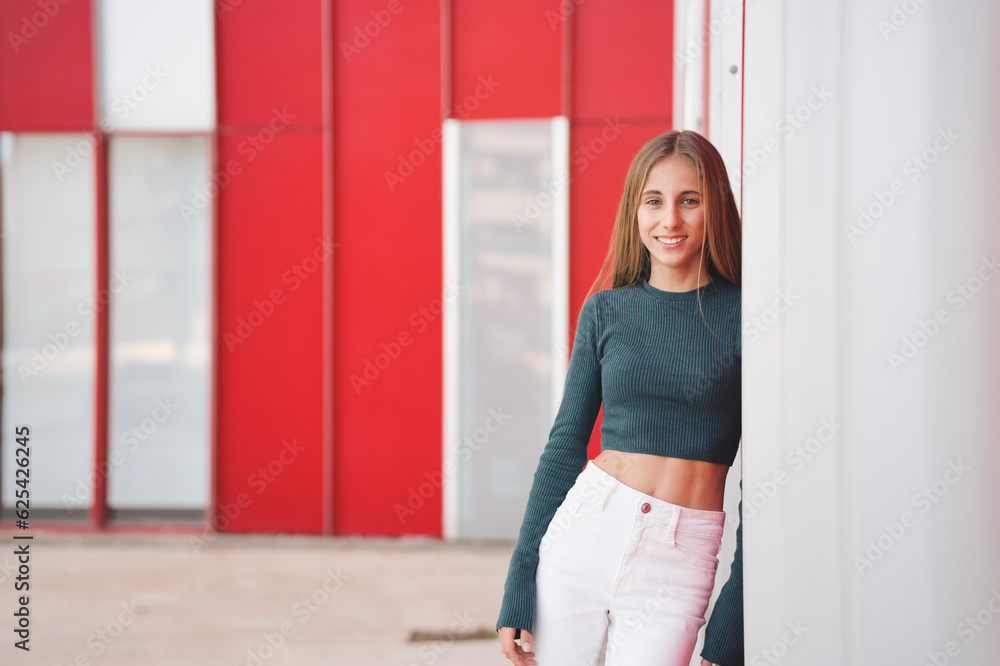 Teenage student, blonde with long hair. Wearing a green sweater and white pants, posing leaning against the wall and looking at the camera. Standing in front of an urban building.