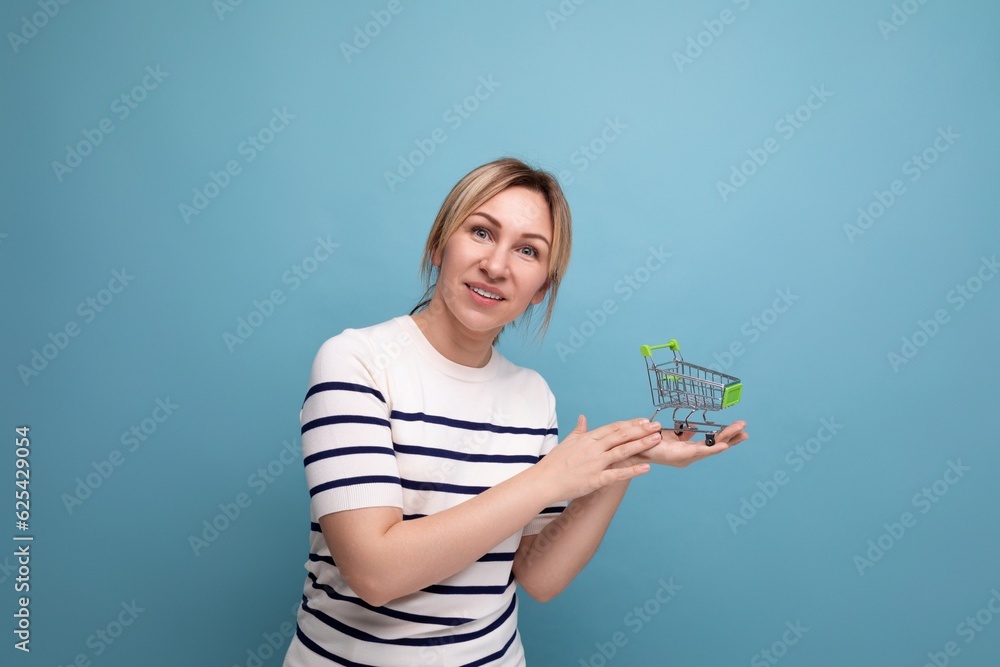 horizontal photo of young cute casual shopaholic woman holding supermarket shopping trolley on blue background with copy space horizontal photo of young cute casual shopaholic woman holding supermarket shopping trolley on blue background with copy space