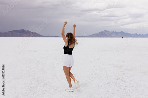 A beautiful unusual landscape on which a large desert of white salt and blue mountains in the distance on a sunny clear day. A young woman in white shorts and black top walks in desert of white salt.  photo