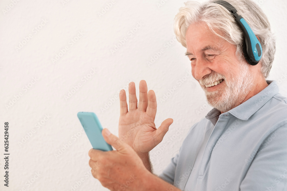 Smiling senior man wear earphones wave to camera having video call on mobile phone. Happy elderly bearded male in headphones on white background talk using modern technologies and wireless connection Smiling senior man wear earphones wave to camera having video call on mobile phone. Happy elderly bearded male in headphones on white background talk using modern technologies and wireless connection