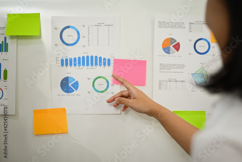 Female economist analyzing sales statistics document, discussing information on whiteboard with her colleague