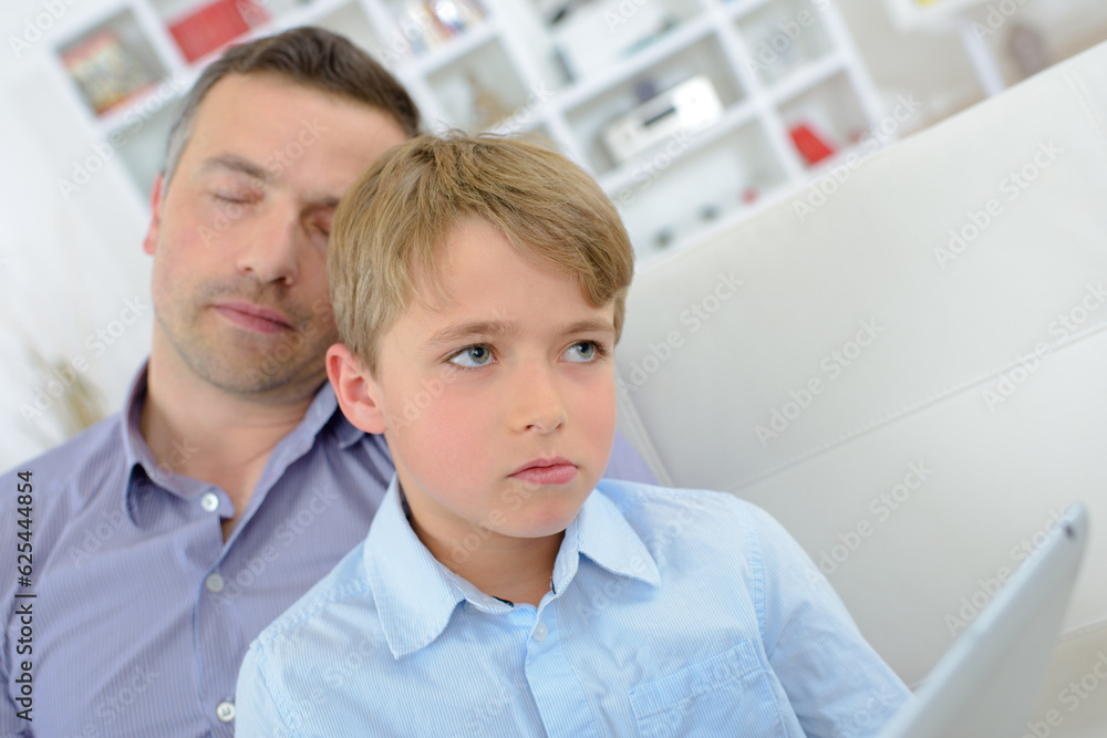Boy on tablet looking pensive while father sleeps Boy on tablet looking pensive while father sleeps