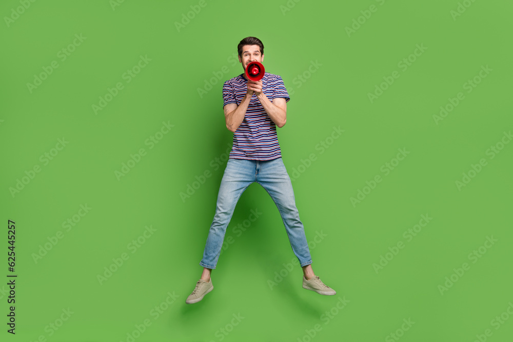Full length photo of impressed cool man dressed striped t-shirt jumping high shouting load speaker isolated green color background Full length photo of impressed cool man dressed striped t-shirt jumping high shouting load speaker isolated green color background
