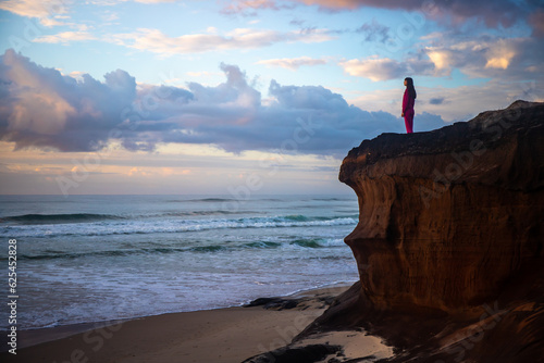 A beautiful girl in a pink tracksuit stands on a rock on Ten Mile Beach and enjoys a stunning orange sunrise over the Pacific ocean. Black rocks campsite, NSW, Australia 