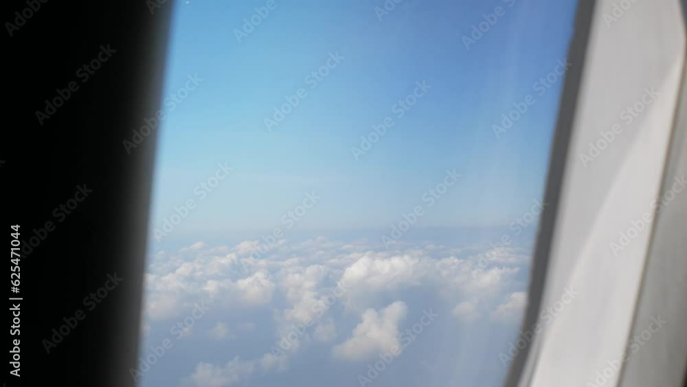 Aerial view through plane window with view over puffy white cloud with clear blue sky while flying, View from the window of the plane traveling by air Aerial view through plane window with view over puffy white cloud with clear blue sky while flying, View from the window of the plane traveling by air