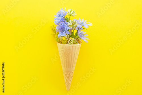 Chicory flowers and other wild flowers in an ice cream cone on a yellow background. Flat top view. © Ann Stryzhekin