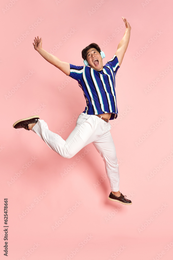 Full-length portrait of young emotional man listening to music in headphones and jumping against pink studio background. Concept of human emotions, lifestyle, casual fashion, positivity, ad