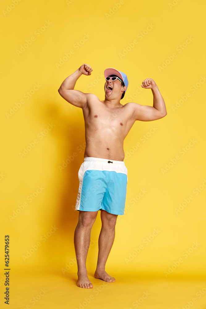 Full-length portrait of positive and excited young man in swimming trunks, sunglasses and cap posing against yellow studio background. Concept of summer, vacation, leisure time, holidays, emotions, ad