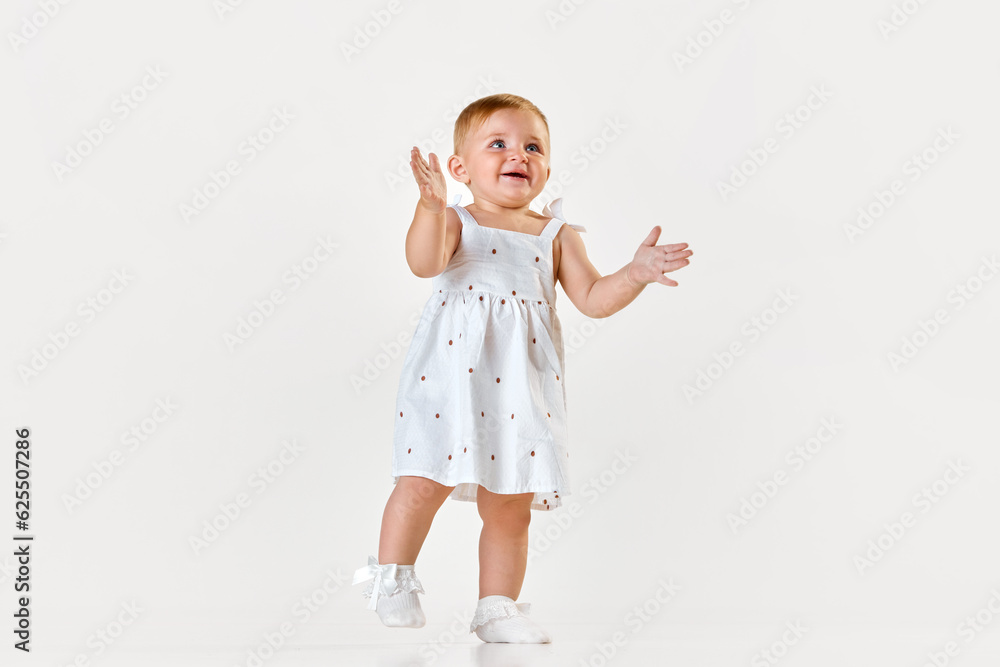 Portrait of little baby girl, toddler in beautiful dress walking, smiling and clapping hands over white studio background. Concept of childhood, newborn lifestyle, happiness, care. Copy space for ad Portrait of little baby girl, toddler in beautiful dress walking, smiling and clapping hands over white studio background. Concept of childhood, newborn lifestyle, happiness, care. Copy space for ad