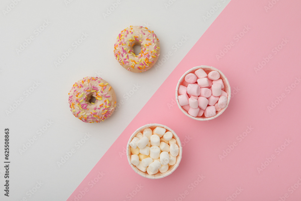 Donuts and marshmallows in paper cups on pink and white background, top view